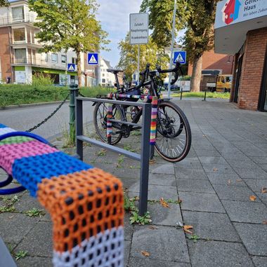 Fahrradständer eckiger Rahmen mit bunten Garn umhäkelt, im Hintergrund weitere Ständer un ein Fahrrad